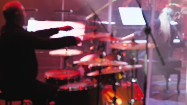 A drummer playing on a drum kit. A man in black clothes behind drums. The end of composition musician beats on different cymbals. Red stage lighting. A bright light and smoke show. Blurred video