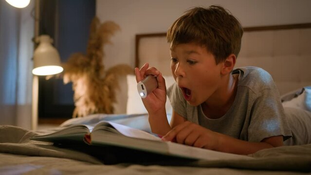 Surprised amazed smiling funny Caucasian child kid boy schoolboy reading interesting book evening in bedroom using flashlight lying on bed home enjoying read fairytale amazing story for night bedtime