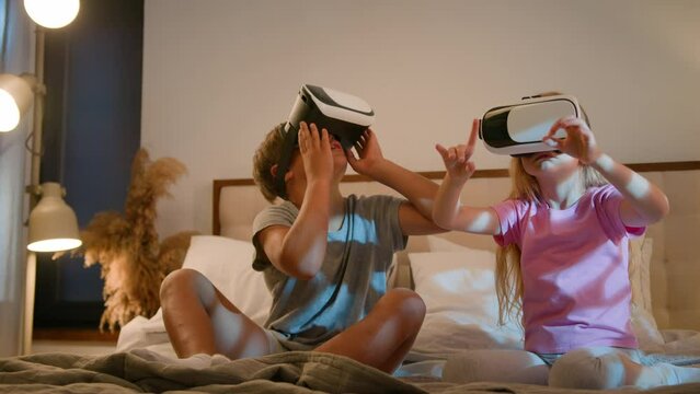 Funny little amazed caucasian children kids boy girl brother sister playing video game using digital virtual reality headset play online metaverse 3d with VR glasses in bedroom innovative technology