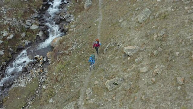 An amazing hike along a stormy mountain cold river. Mountain tourists with large backpacks and trekking poles walk along a trampled narrow path. Ala-Archa National Park in Kyrgyzstan. Active leisure.