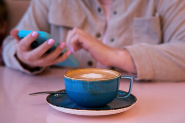 Unrecognizable Woman Using Mobile Phone and Drinking Coffee in a Cafe, Browsing Social Media. Female Person Sitting in a Coffee Shop with Blue Ceramic Cup of Cappuccino. Hands with Smartphone Close up