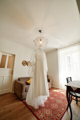 Fototapeta na wymiar White bride dress with a long train hangs on a hanger on a chandelier in the room