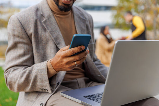 Businessman Sitting With Laptop And Using Smart Phone At Office Park