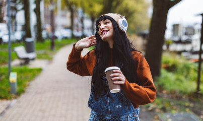 Happy beautiful woman holding coffee cup and enjoying music through wireless headphones on footpath