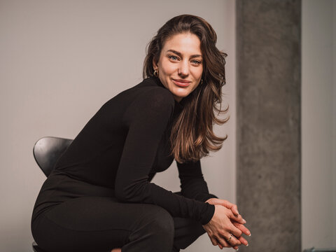 Smiling Woman Sitting On Chair With Hands Clasped In Front Of Wall