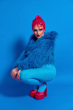 Non binary person with pink hair crouching against blue background