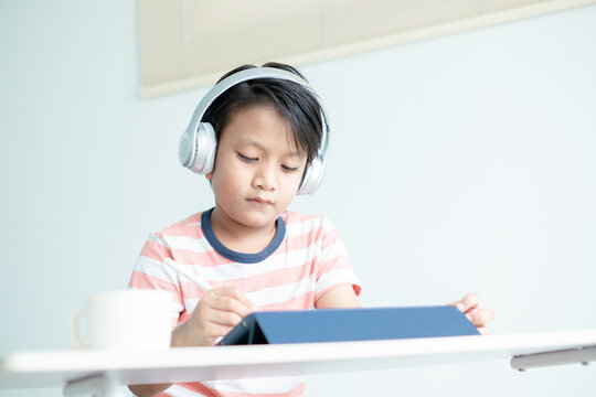 Asian boy kid with headphones is drawing pictures and writing on the digital tablet using digital pencil