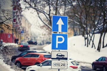 Paid parking lots in downtown, park sign on pole in winter day. Row of cars parked on snowy roadside in winter, paid parking sign. Winter parking problems.