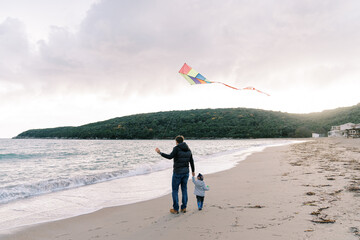 Dad with a kite on a string stands with a little girl on the seashore. Back view