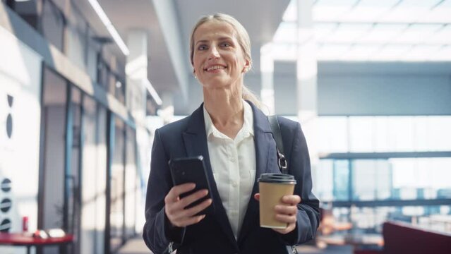 Medium Shot of Business Woman in a Tailored Suit Walking in a Business Office, checking her Smartphone. Confident Woman on Her Way to Do Important Deals with Investors, Preparing Notes for a Meeting