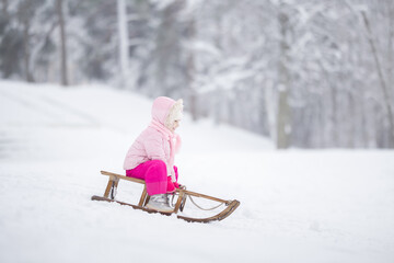 Happy little girl in pink warm clothes sitting on sledge and sledding down on snow from hill. Child...