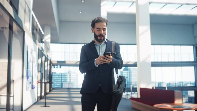 Young Fun and Handsome Businessman Celebrates Through Open Space Office Hallway, Using Smartphone. Male Latin Worker in a Suit Celebrating his Latest Achievement, Receiving Happy News of Promotion