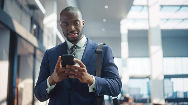 Slow Motion Portrait of Handsome Black Businessman in Stylish Suit Standing In Office Building. Smiling Male CEO Using Smartphone To Check Work Emails from Employees and Investors