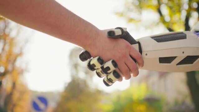 Shot of robot shaking human hand on blurred background of autumn park in sunlight outdoor. Unrecognizable person. Artificial intelligence concept