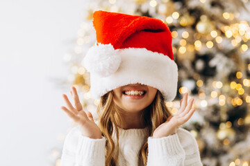 Portrait of cute girl standing in front of Christmas tree wearing santa hat and smiling