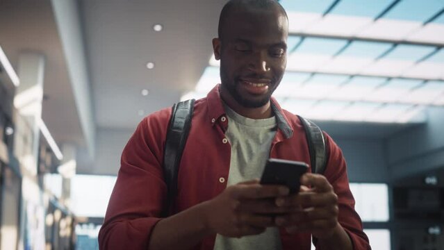 Portrait of a Young Black Man Walking Across Corporate Office Hall, Browsing Web on his Smartphone and Smiling. Successful Entrepreneur Walks and Thinks of Startup Strategy. Low Angle