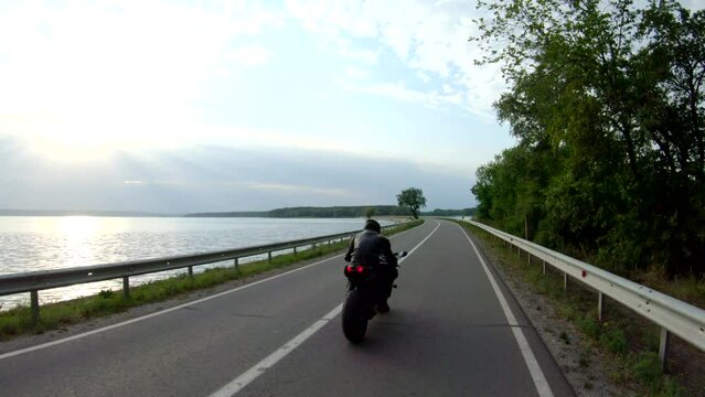 Biker riding on modern sport motorbike through road near lake. Motorcyclist racing motorcycle at dam route on summer day. Man driving bike through bridge of river. Concept of freedom and lifestyle