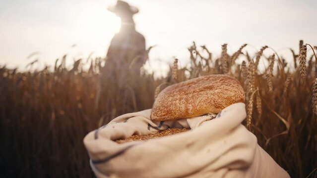 Wheat grain bag with loaf bread on field. Unrecognizable farmer in straw hat on agricultural wheat field at sunset on background. Grain, food production, agribusiness harvesting summer season concept.