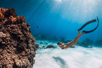 Woman free diver in bikini swims underwater with fins near corals in tropical blue sea