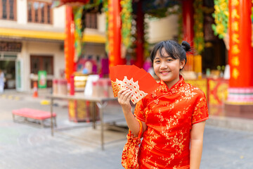 Chinese lunar new year festival and tradition holiday celebration concept. Happy Little Asian girl in Chinese red dress holding money gift in red envelopes in front of in Chinese temple shrine.