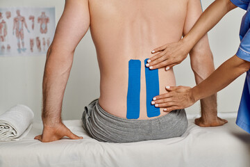 cropped view of young female doctor putting blue kinesio tapes on back of her patient, healthcare