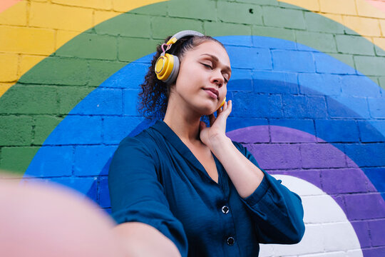 Young woman wearing wireless headphones taking selfie with eyes closed in front of rainbow wall