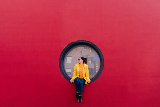 Woman in yellow rain coat sitting in porthole in red wall