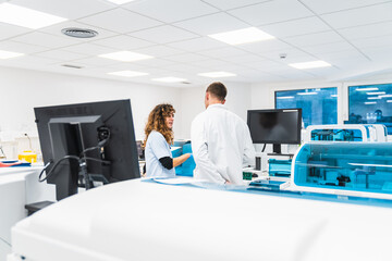 Two lab technicians converse amidst blood analysis machines in a clean, bright hospital laboratory setting.