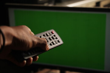 man's hand holding a tv remote with an empty green screen monitor background