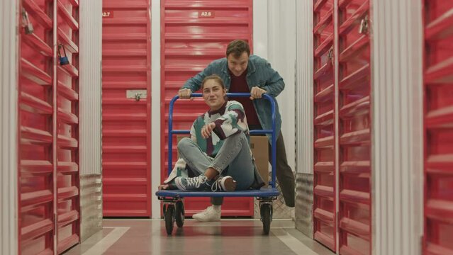 Slow motion shot of cheerful man running through corridor and pushing excited wife on dolly cart while having fun in self service warehouse
