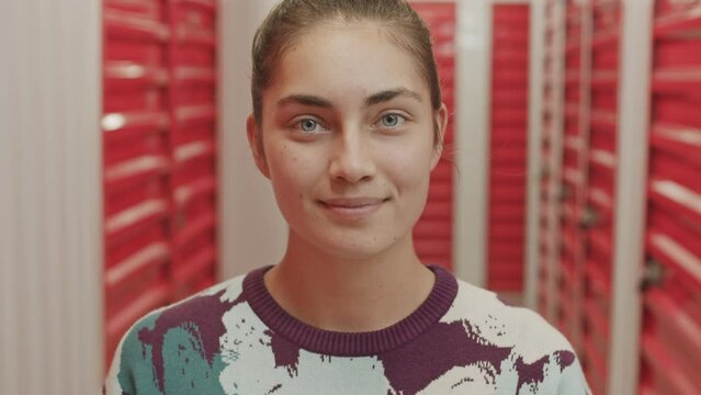 Tilt up portrait shot of young cheerful woman holding box with fragile sticker and smiling on camera while posing in self service storage