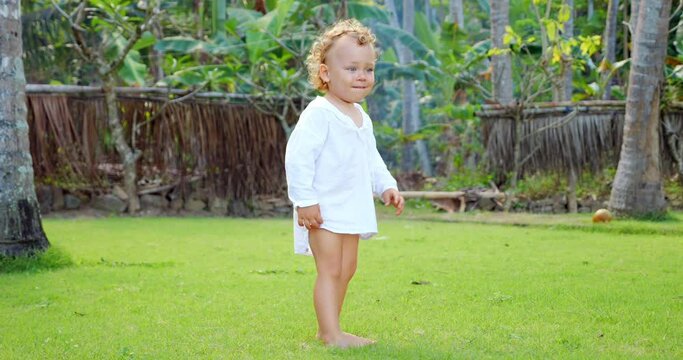 Toddler's emotions shift from perplexity and frustration to carefree mood in this slow-motion, full-length portrait shot outdoors in tropical garden. Short clip of young child wearing light white