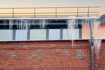 Melting icicles on roof and drain pipe of the building. Icicles hanging on roof and gutter of modern building. Sharp icicles hang from edge of the roof. Icicles above the entrance, risk to life - obrazy, fototapety, plakaty