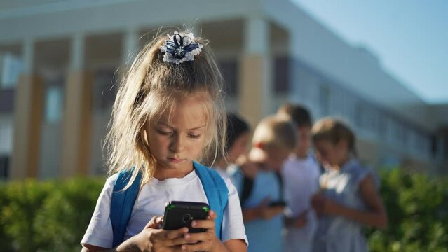 Children stand in schoolyard and look at smartphone.students with backpacks at school.children play smartphones in school yard after school.students listen to music on smartphone.happy family concept