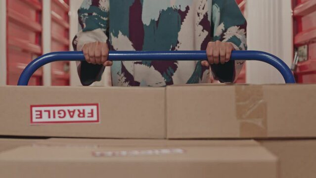 Close up shot of unrecognizable woman pushing dolly cart with boxes marked as fragile while walking through self storage corridor