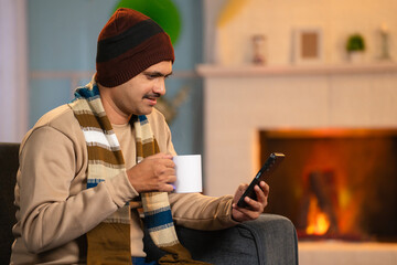 Indian middle aged man in winter wear using mobile phone while drinking tea or coffee at home -...