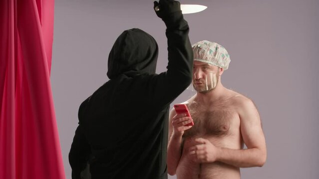 A maniac in a mask and with a knife pulls back the shower curtain, wanting to attack a man. A man in a cap with a hydrogel mask on his face and a smartphone looks at the maniac with a bored look.
