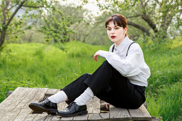 A beautiful girl in a white shirt, in black trousers with suspenders against the background of the sky and green grass