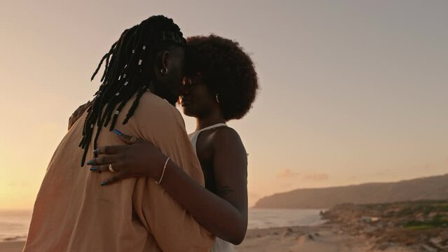 Concept of Love and passion, a stunning black woman in a white dress embraces a handsome African man with dreadlocks on the beach at sunset, sharing a moment of intense connection