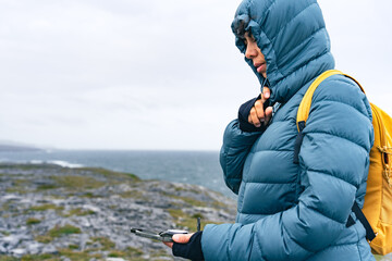 Latin woman sheltering from the wind with a hood while looking at the compass in a landscape with the sea in the background on the Irish coast - obrazy, fototapety, plakaty