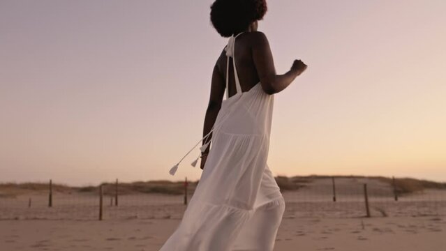 A stunning black woman in a fashionable white dress gracefully running on the sand at sunset, embodying elegance and beauty in motion