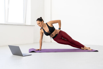 Young woman in sportswear planking on yoga mat, exercising at home, using laptop, watching fitness video