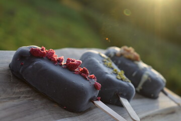 Ice cream popsicle covered in chocolate with walnut, pistachio and raspberry flavor on wooden board in sunlight
