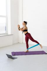 Young woman with resistance band in the morning at home and using online lesson on laptop