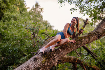 Fit girl exercises outdoors in a green park, climbing trees for strength and flexibility. Enjoying nature, she warms up before her workout, embracing a healthy lifestyle.