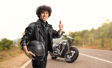 Young man in a black leather jacket gesturing thumbs up in front of a motorbike