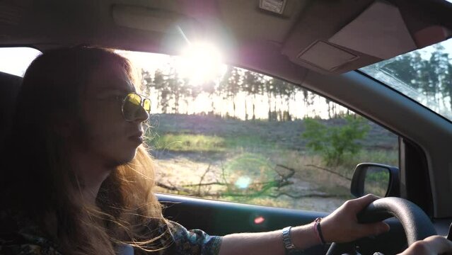 Profile of hippie man in sunglasses driving a motor car with sunlight at background. Young male hipster with long hair riding on auto to rural road. Concept of journey and trip. Close up Slow motion