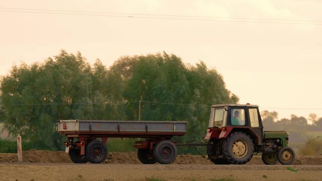 Farmer in vintage old tractor with trailer is driving along road along fields. Concept of agronomy and farming. Tractor transporting corn or wheat cargo from field during harvesting