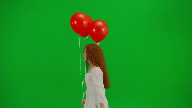 Redheaded little girl in white dress running with red balloons on green background of studio. Side view. Concept of holiday, joy and fun.