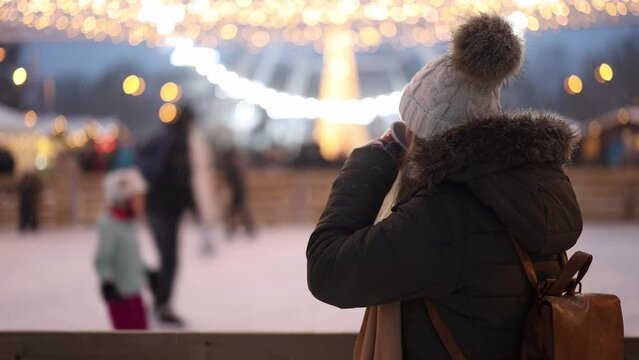 Woman looks at people skating on ice rink and drinks mulled wine or punch. Christmas market and winter holidays in city